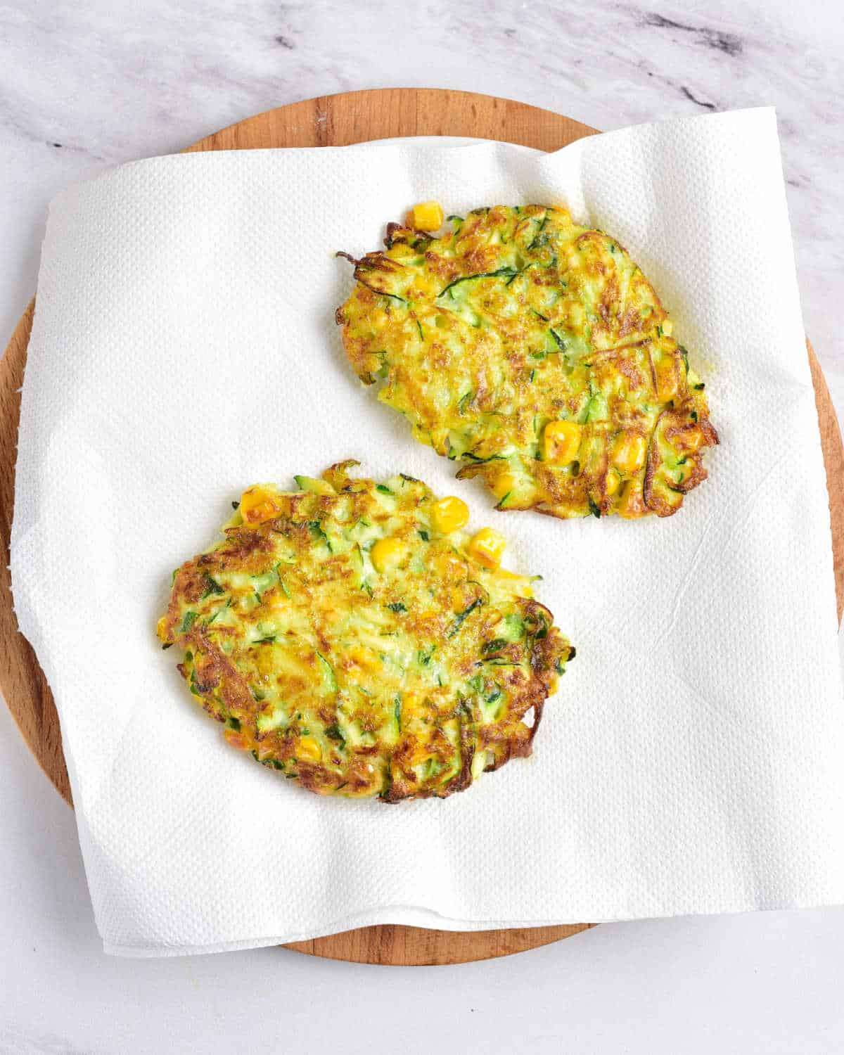 Blotting two corn zucchini fritters on white paper on a board. Grey marbled surface. 