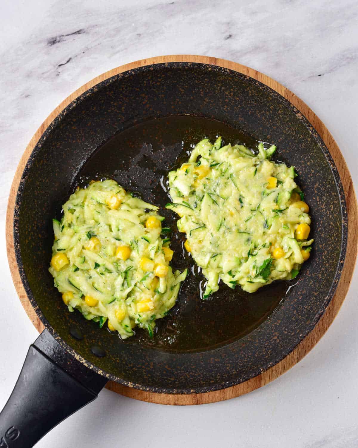 Grey marbled surface with dark skillet containing two zucchini corn fritters before flipping.