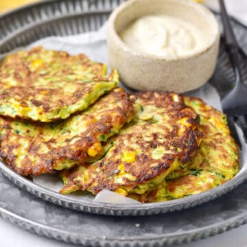Overlapping zucchini corn fritters on a metal plate with bowl of mayo.