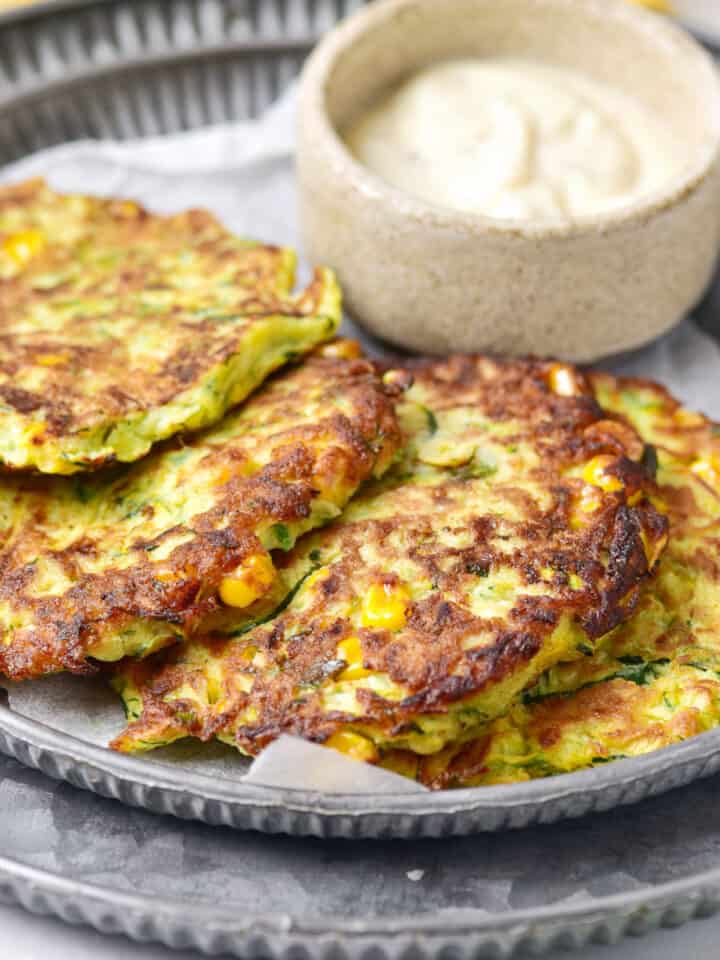 Overlapping zucchini corn fritters on a metal plate with bowl of mayo.