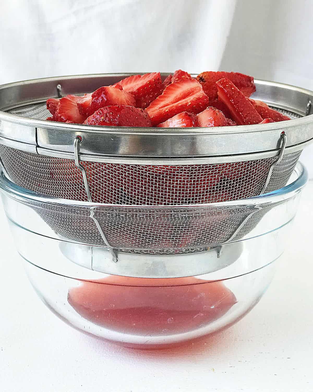 Metal colander draining strawberries over a glass bowl. White background.