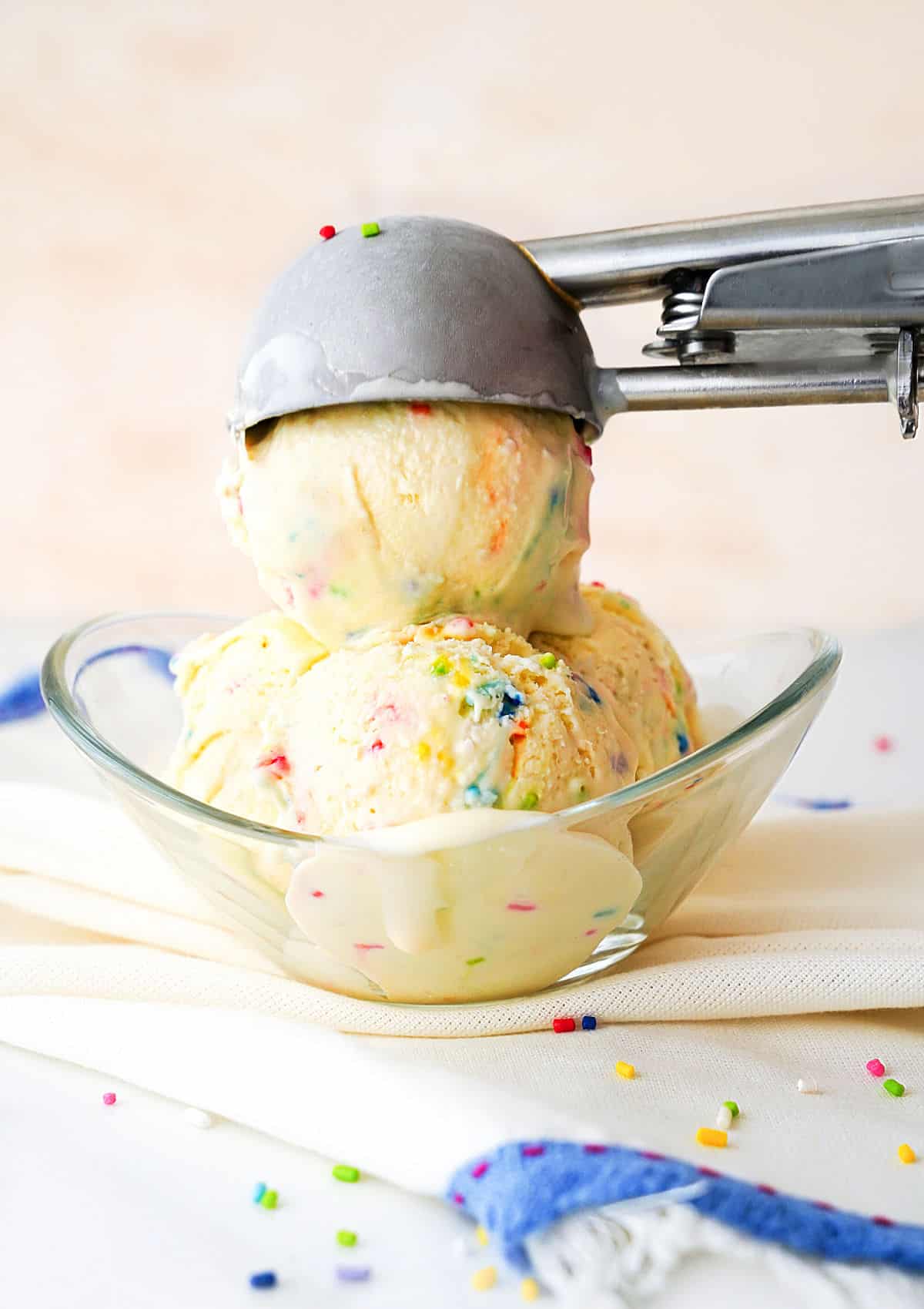 Scooping funfetti ice cream balls in a glass bowl. Pink background white blue striped cloth. 