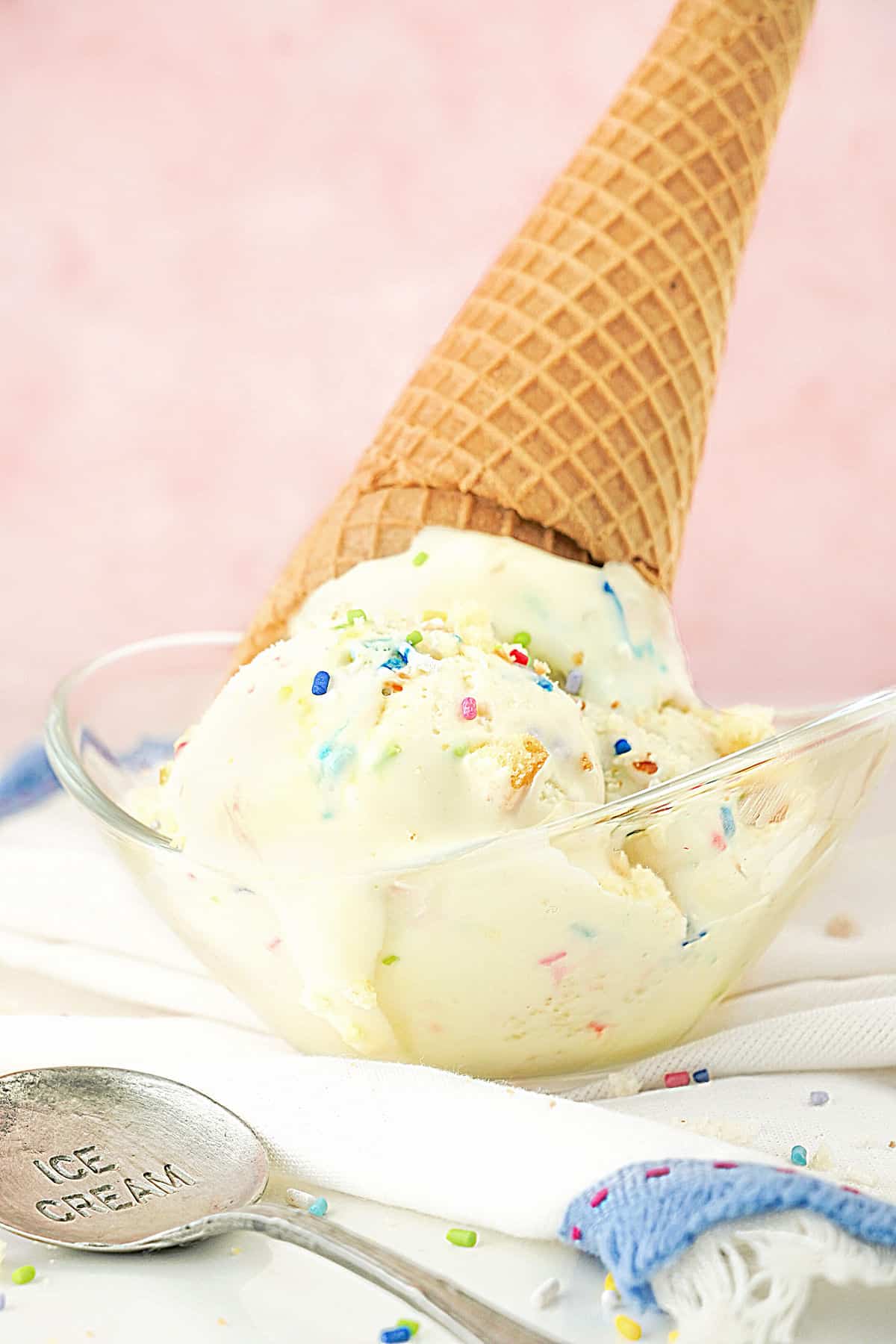 Glass bowl with funfetti ice cream scoops and a waffle cone. Pink background, white cloth, vintage spoon. 