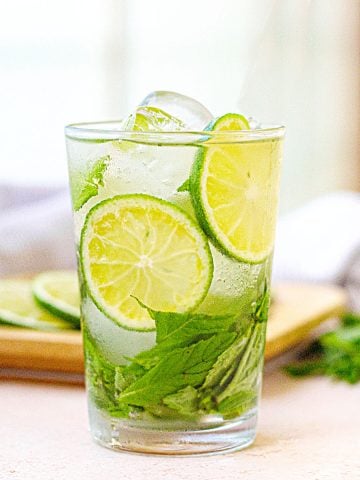 Cucumber mint lime water in a glass on a peach surface with whitish background; a tray with ingredients.