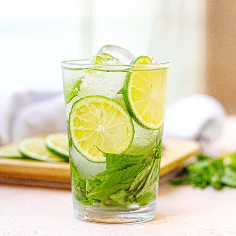 Cucumber mint lime water in a glass on a peach surface with whitish background; a tray with ingredients.