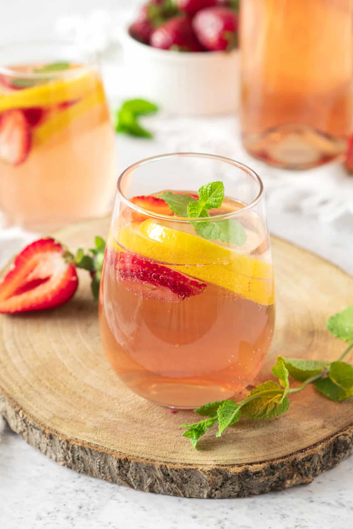 Round wood board with glasses of rose sangria with strawberries and orange slices. Mint leaves, bowl with berries, pitcher. 