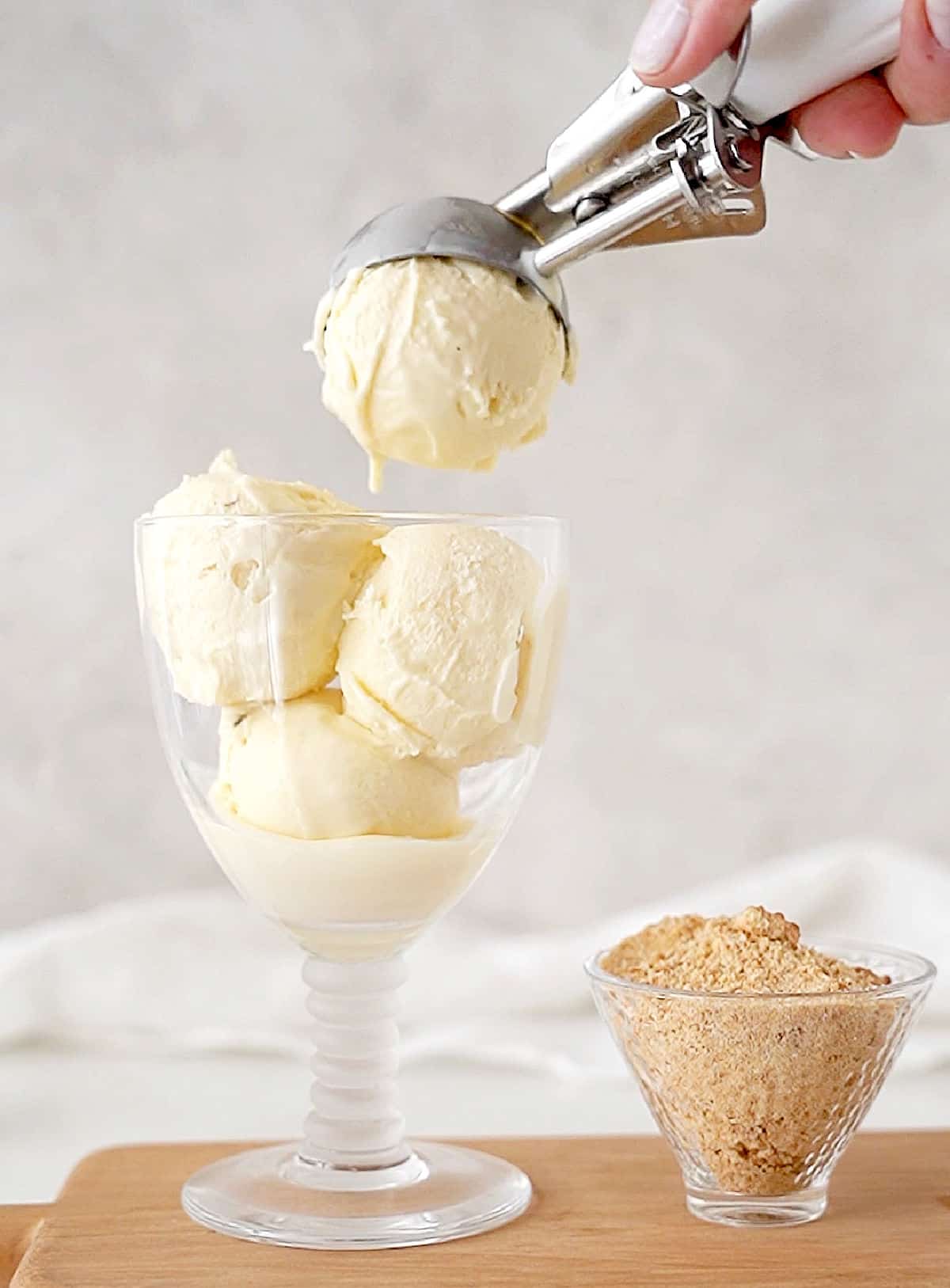 A glass with vanilla ice cream being scooped. Bowl with crumbs, wooden surface, grey background. 