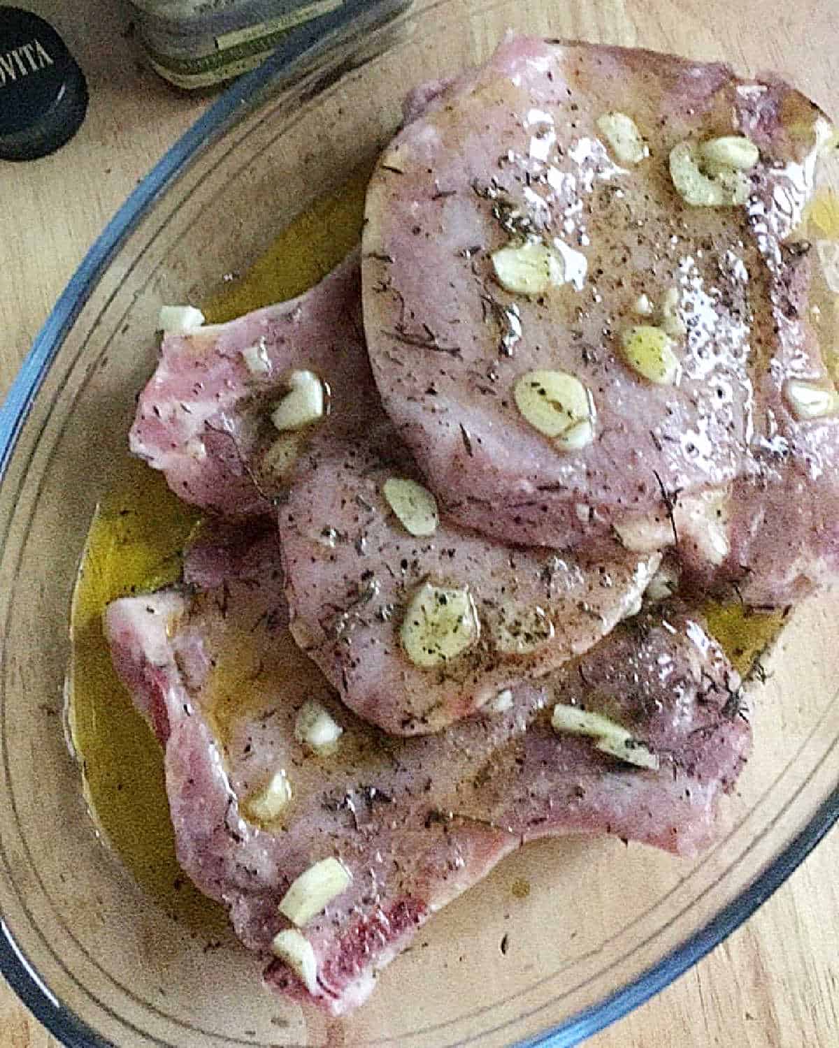 Pork chops marinating with garlic, thyme and olive oil in a glass dish.