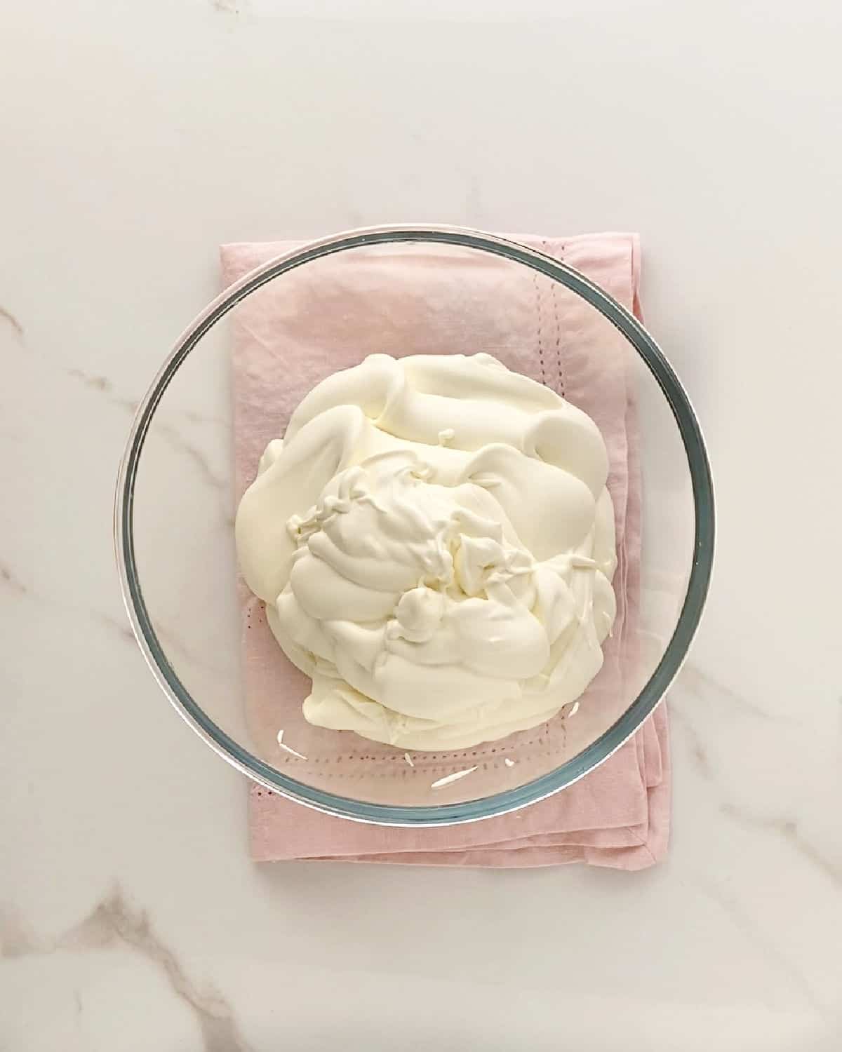 Cream mixture in a glass bowl on a pink napkin. White marble surface.