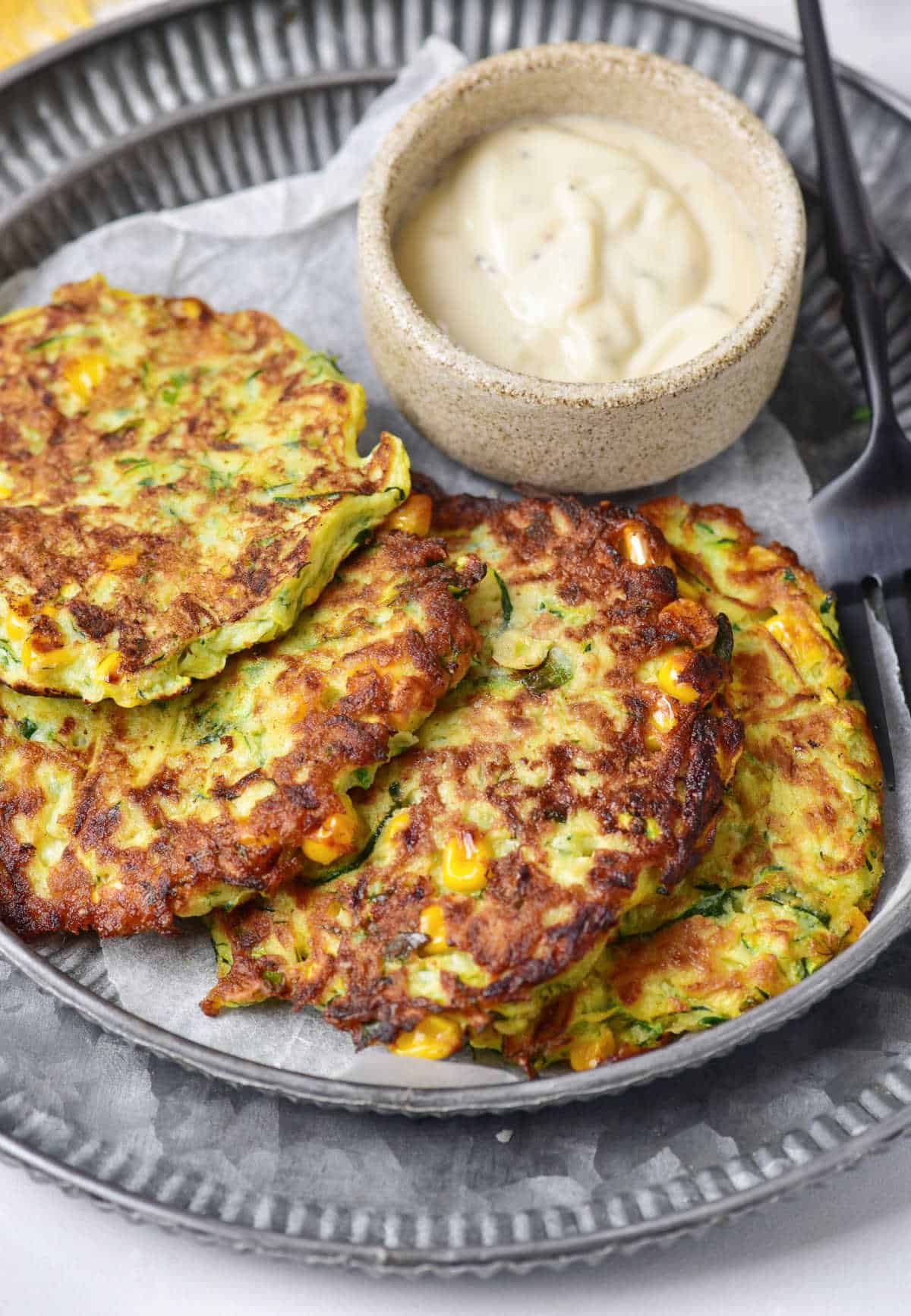 Several zucchini corn fritters overlapping on a dark metal plate. Bowl with mayo.