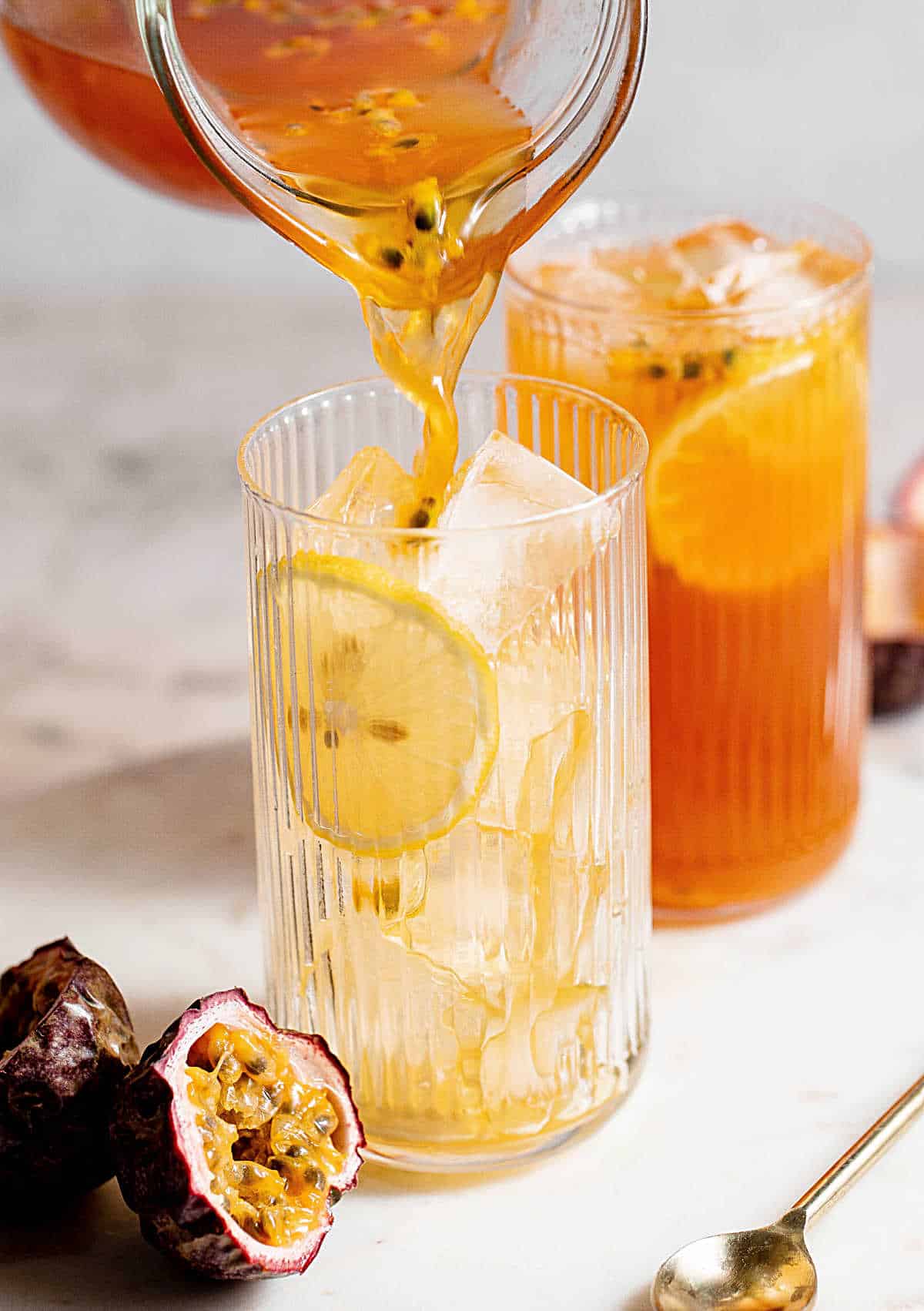 Pouring passionfruit tea from a jug into tall glasses with ice. White marble surface, grey background.