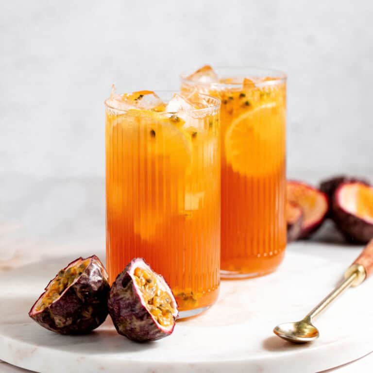 Two tall glasses with passion fruit tea on white marble board. Passionfruit halves, long spoon, grey background.