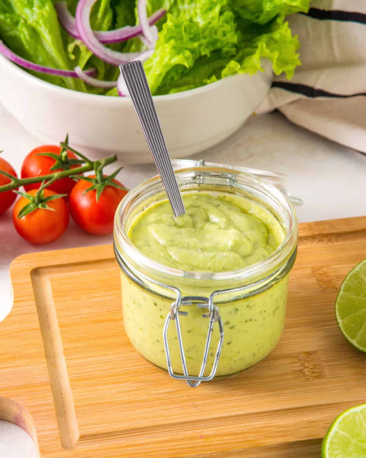 Avocado ranch dressing in a mason jar on a wooden board. Tomatoes, bowl with greens, limes.