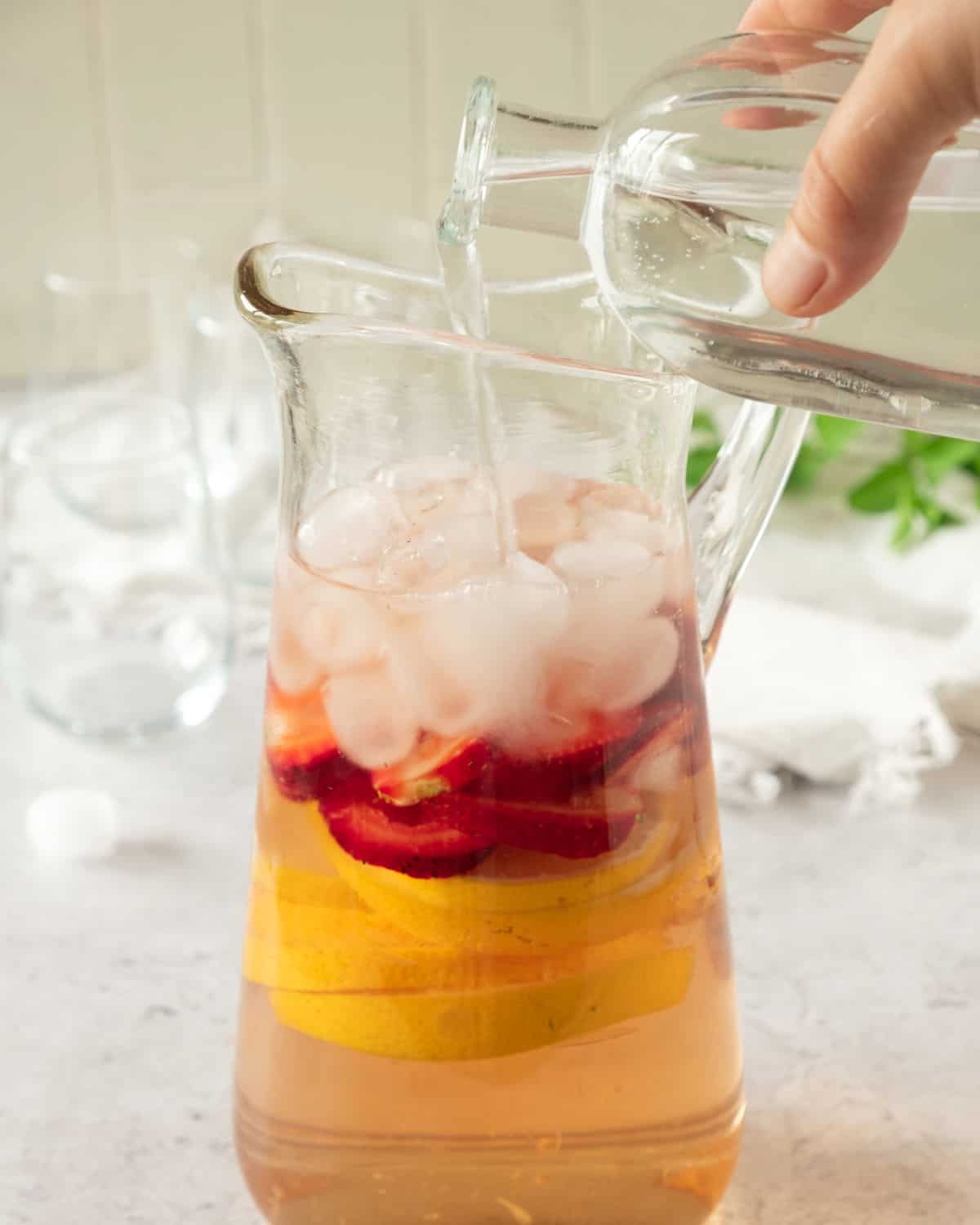 Adding clear liqueur to pitcher with strawberries, orange slices, ice and rose wine. Light grey background with glasses.
