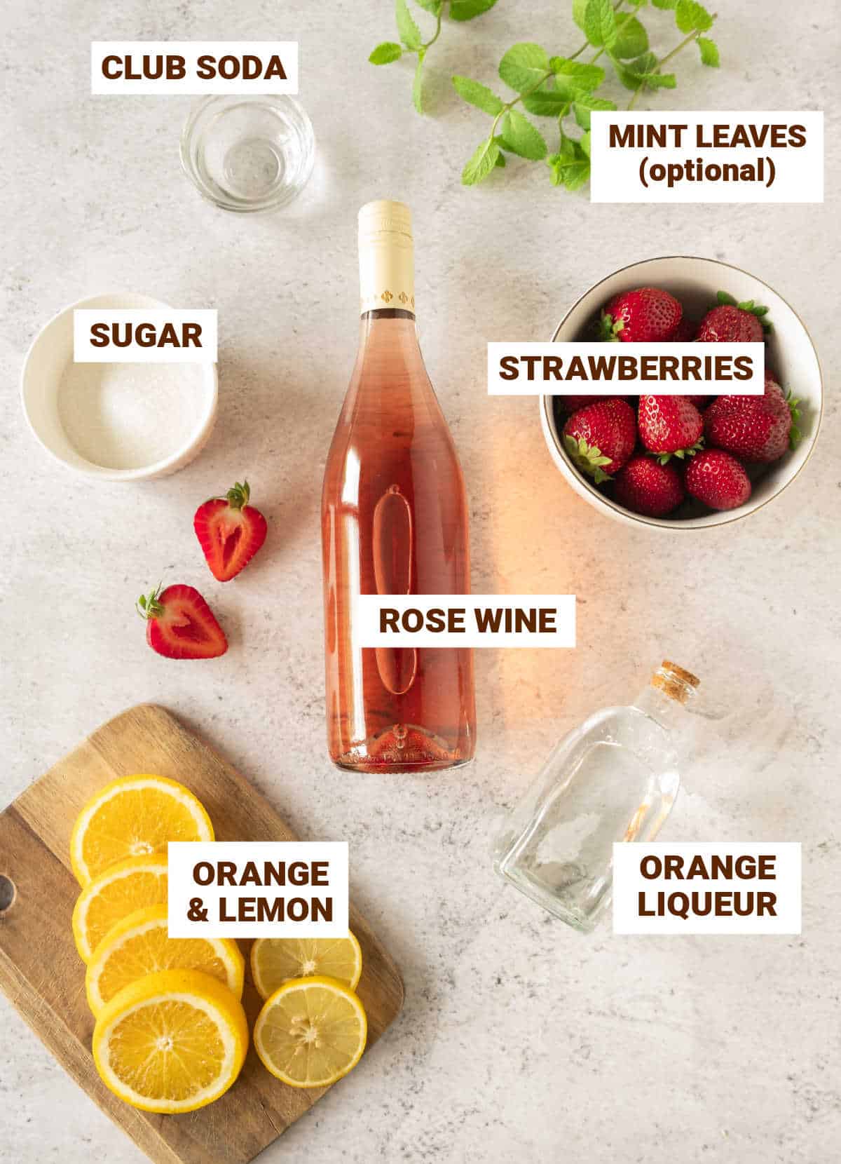 Light grey surface with ingredients for rose wine sangria including citrus slices, strawberries, sugar, mint, liqueur bottle.