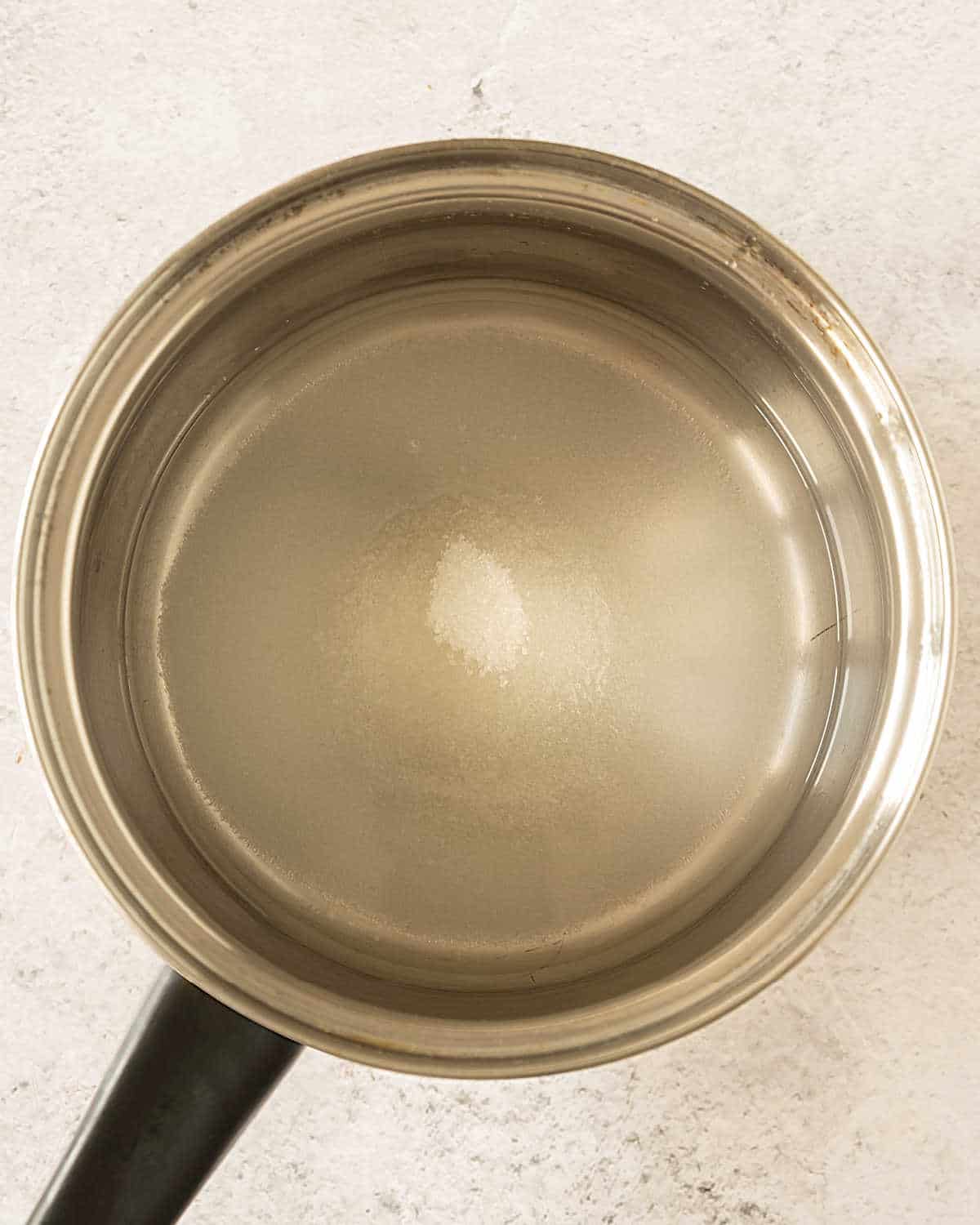 Metal saucepan with sugar and water. Light grey surface.