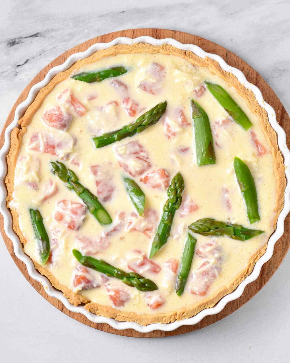 Asparagus pieces top a salmon quiche filling in a round pie dish. White marbled surface. 