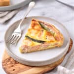 Slice of salmon quiche on a grey plate. Silver fork, wooden board on grey marbled surface.