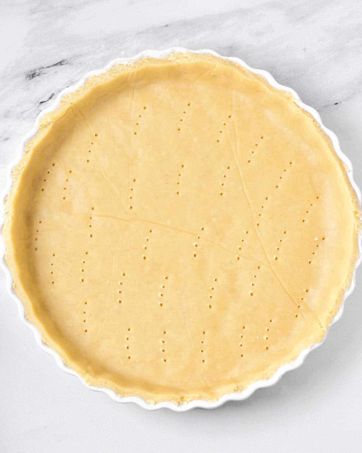 White round dish with unbaked pricked pie dough. White marbled surface.