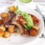 White plate with pork chops, salsa verde and roasted potatoes. A knife, bowl with salsa, white surface.