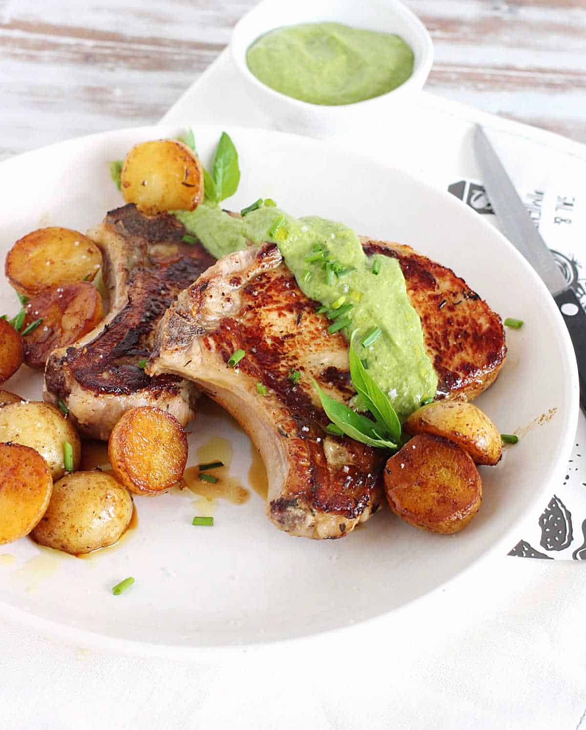 Two pork chops with salsa verde and roasted potatoes on a white plate. Bowl with salsa, whitish surface, a knife.