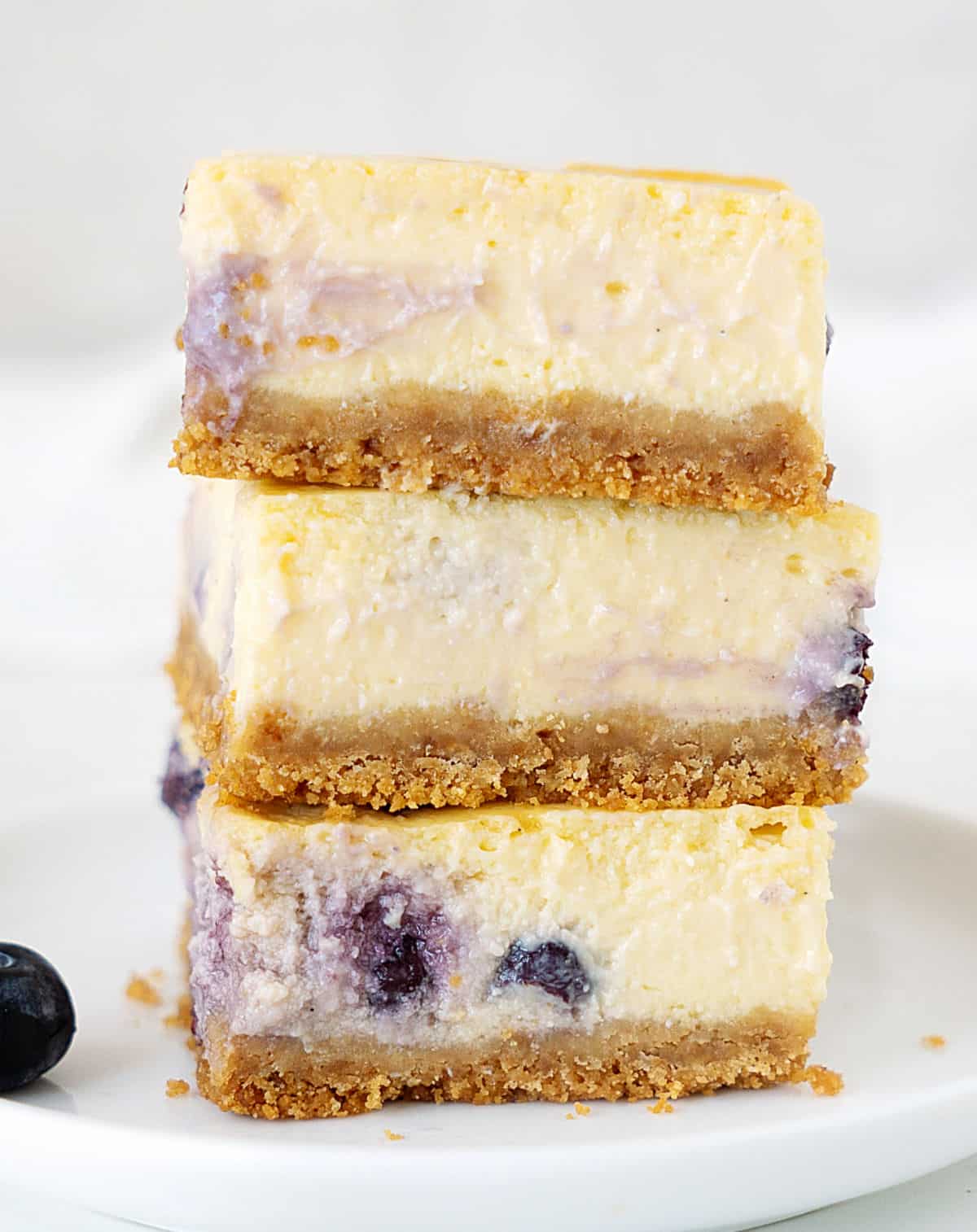 Stack of three blueberry cheesecake squares on a white plate. White grey background.