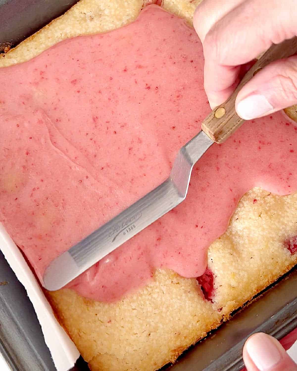 Spreading pink glaze with an offset spatula on top of blondies in a metal pan.
