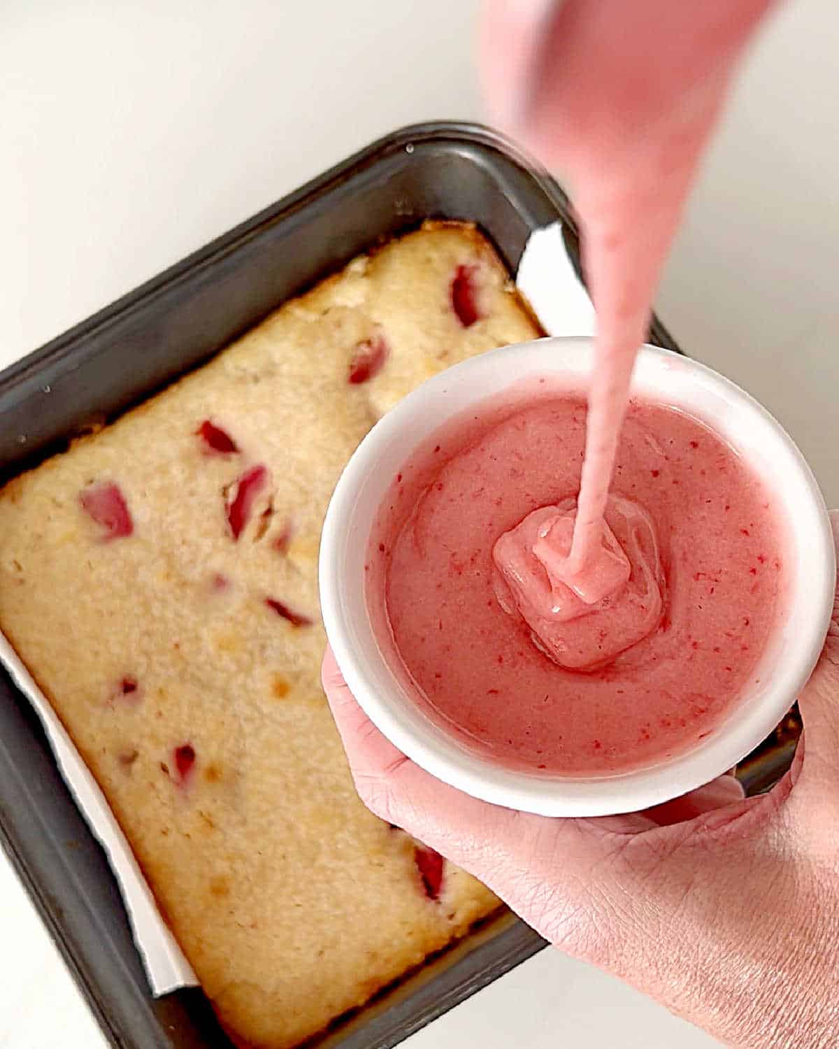 White bowl with strawberry glaze over metal square pan with blondies. White surface. 