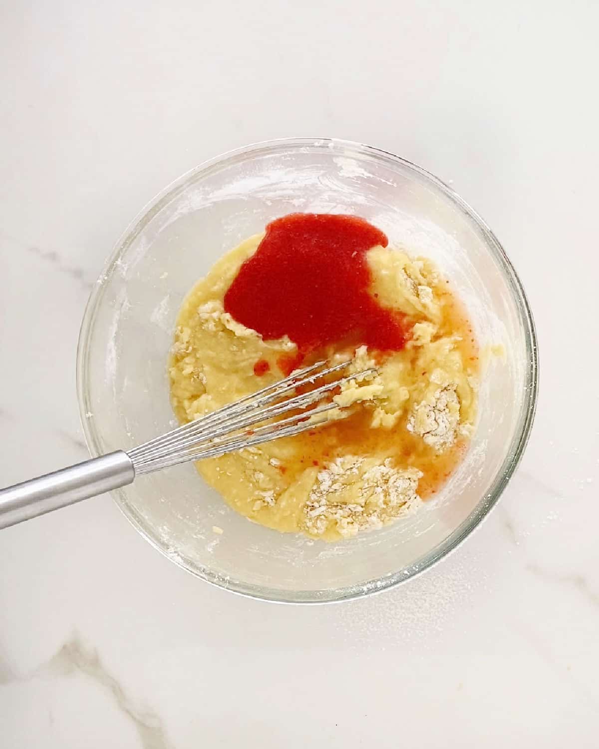 Mixing ingredients for lemon strawberry blondies in a glass bowl with a whisk. White marble surface. 