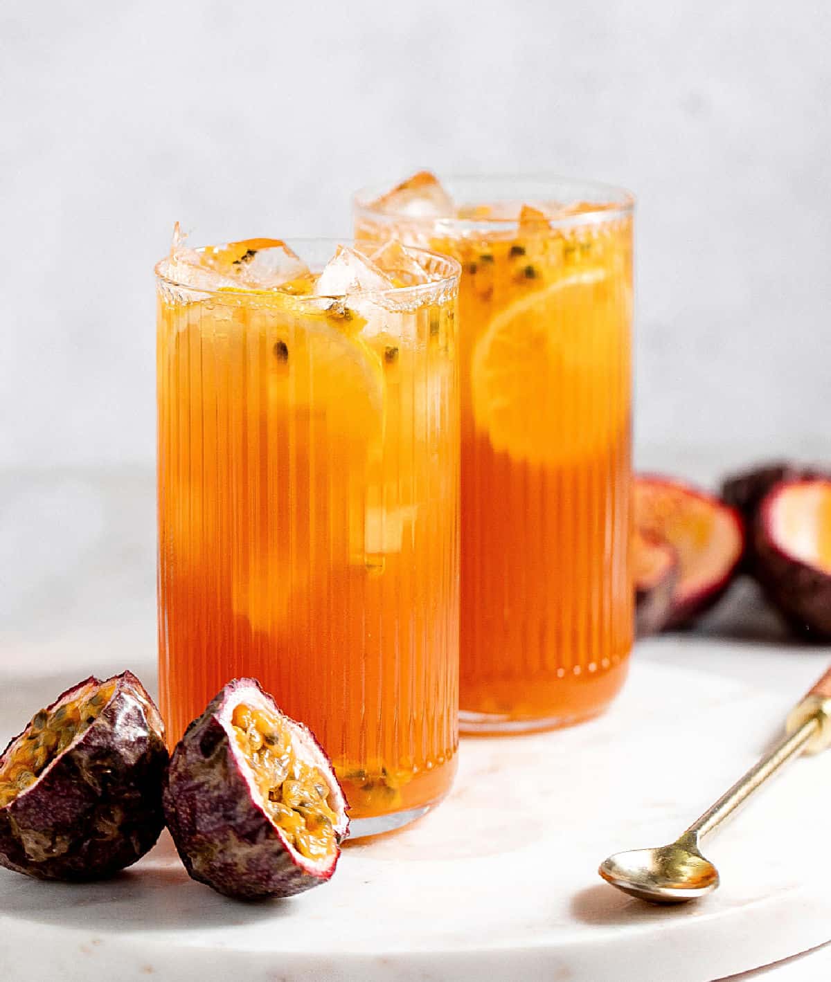 Passionfruit tea served in two tall glasses with grey background and white marble surface. Passion fruits and long spoon.