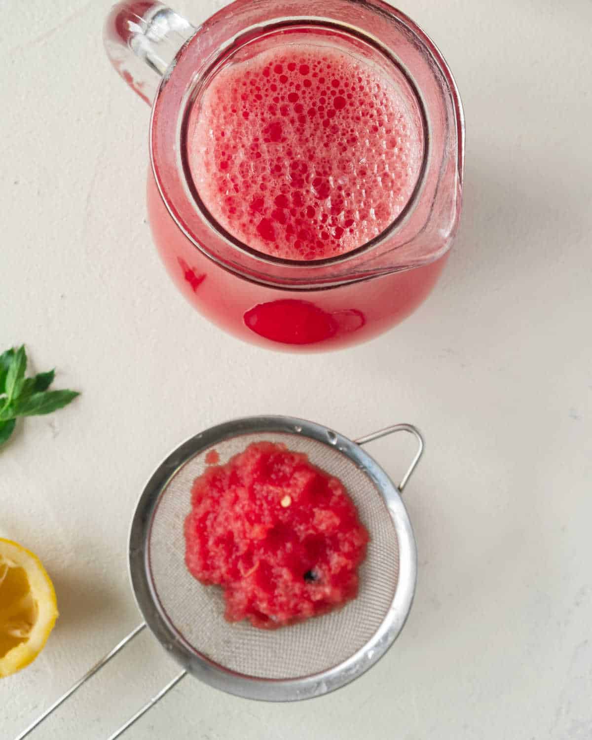 Top view of small jug with watermelon syrup and sieve with pulp. Off-white surface.