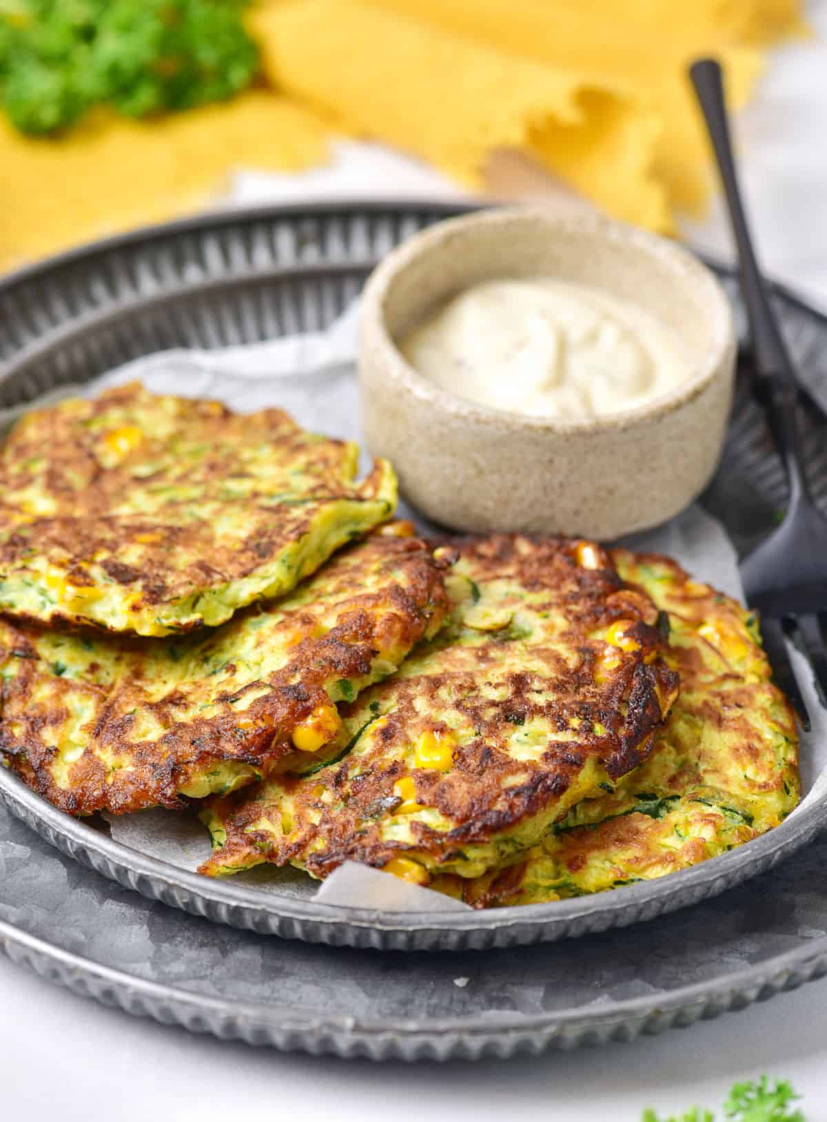 Corn zucchini fritters overlapping on a metal plate. Bowl with mayo. Yellow cloth in the background. 