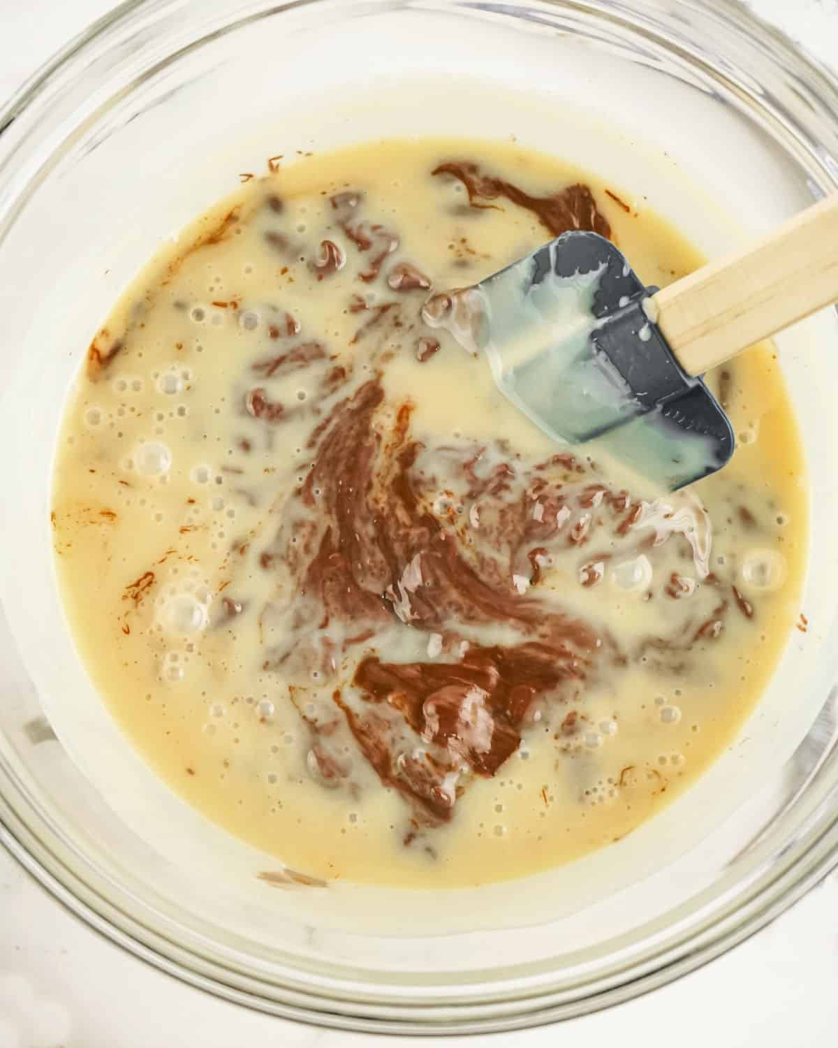Stirring condensed milk with melted chocolate in a glass bowl with a spatula.