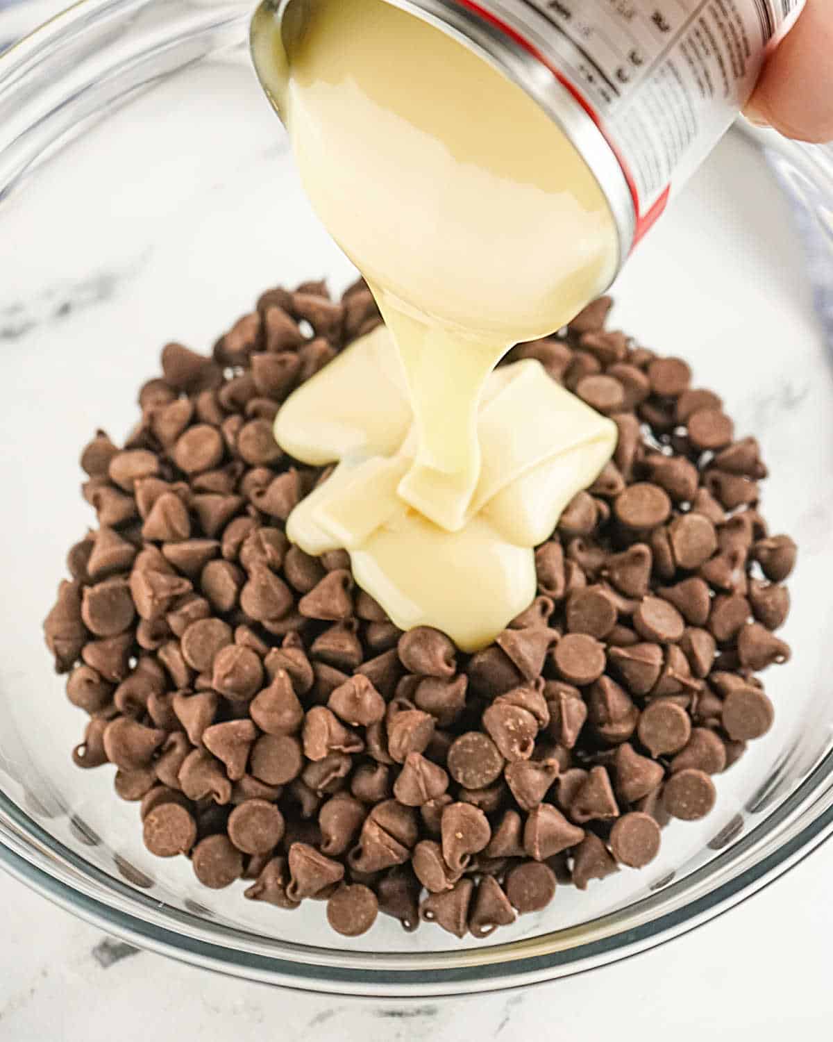 Pouring a can of condensed milk on chocolate chips in a glass bowl.
