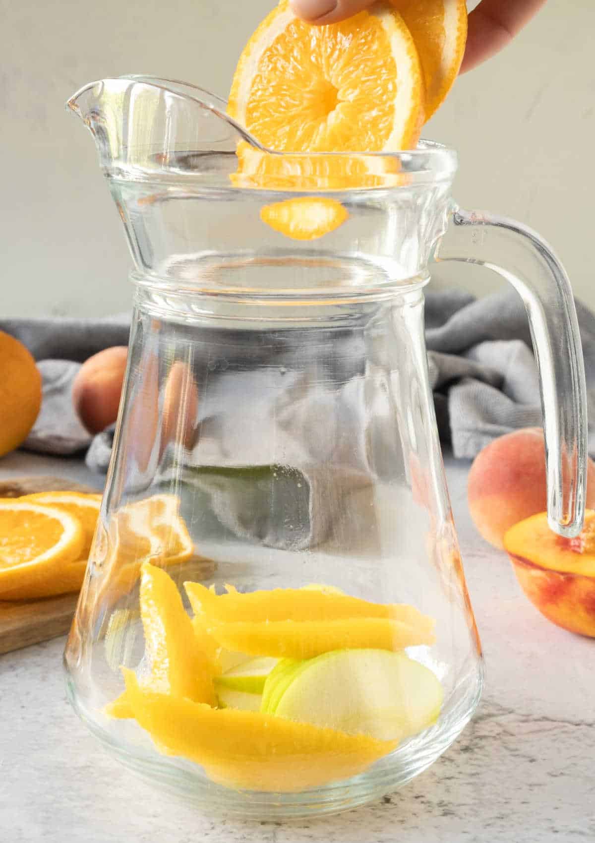 Adding sliced fruit to a large pitcher on a grey surface and beige background.