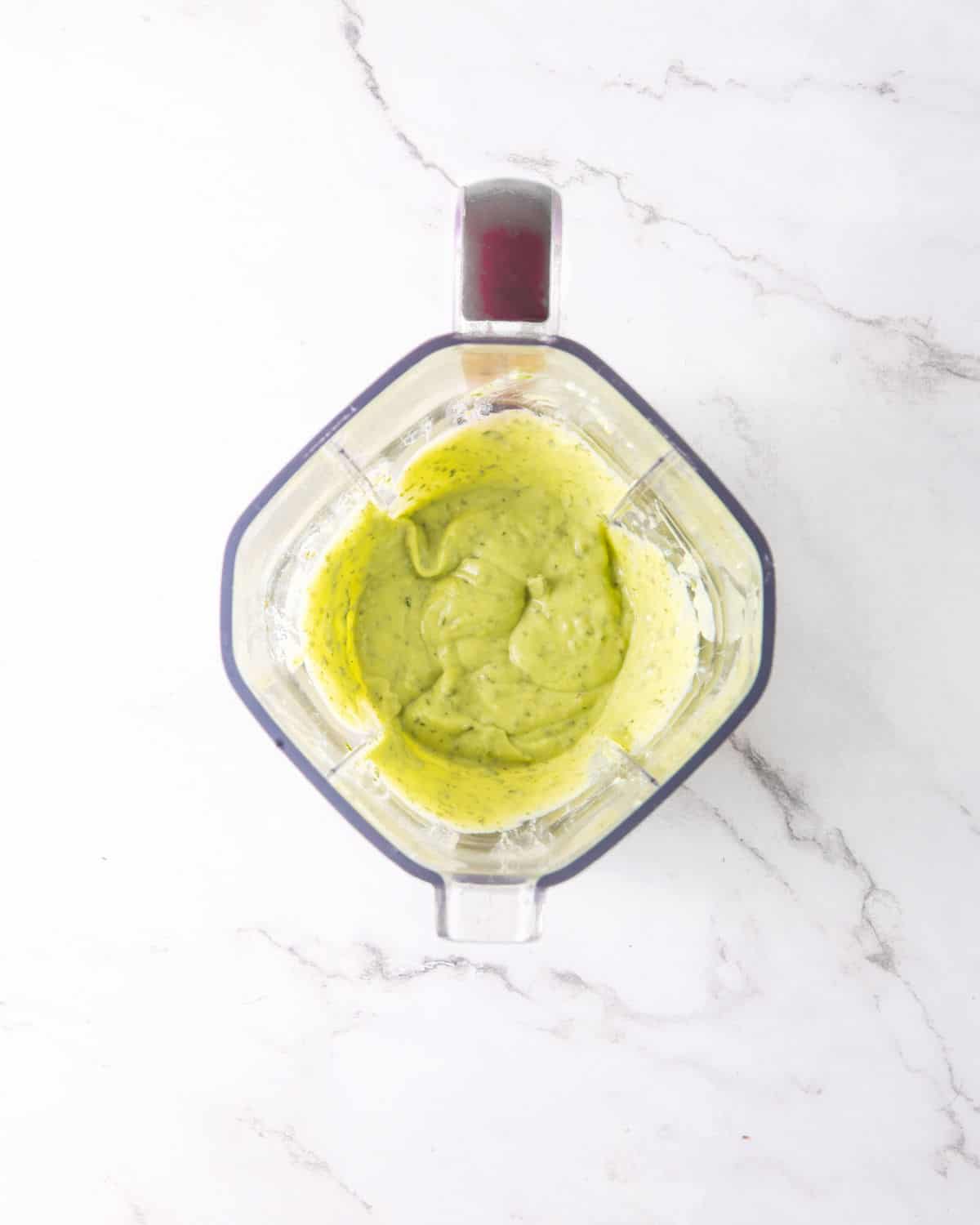 Blended avocado ranch dressing in the jar of a blender on a whit marbled surface.