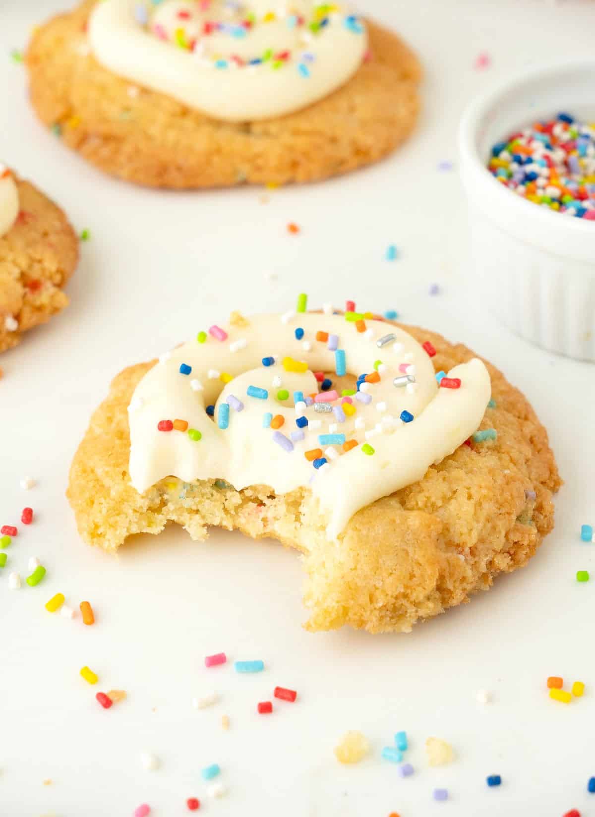 Bitten frosted funfetti cookie on a white surface with sprinkles. Whole cookie in the background.
