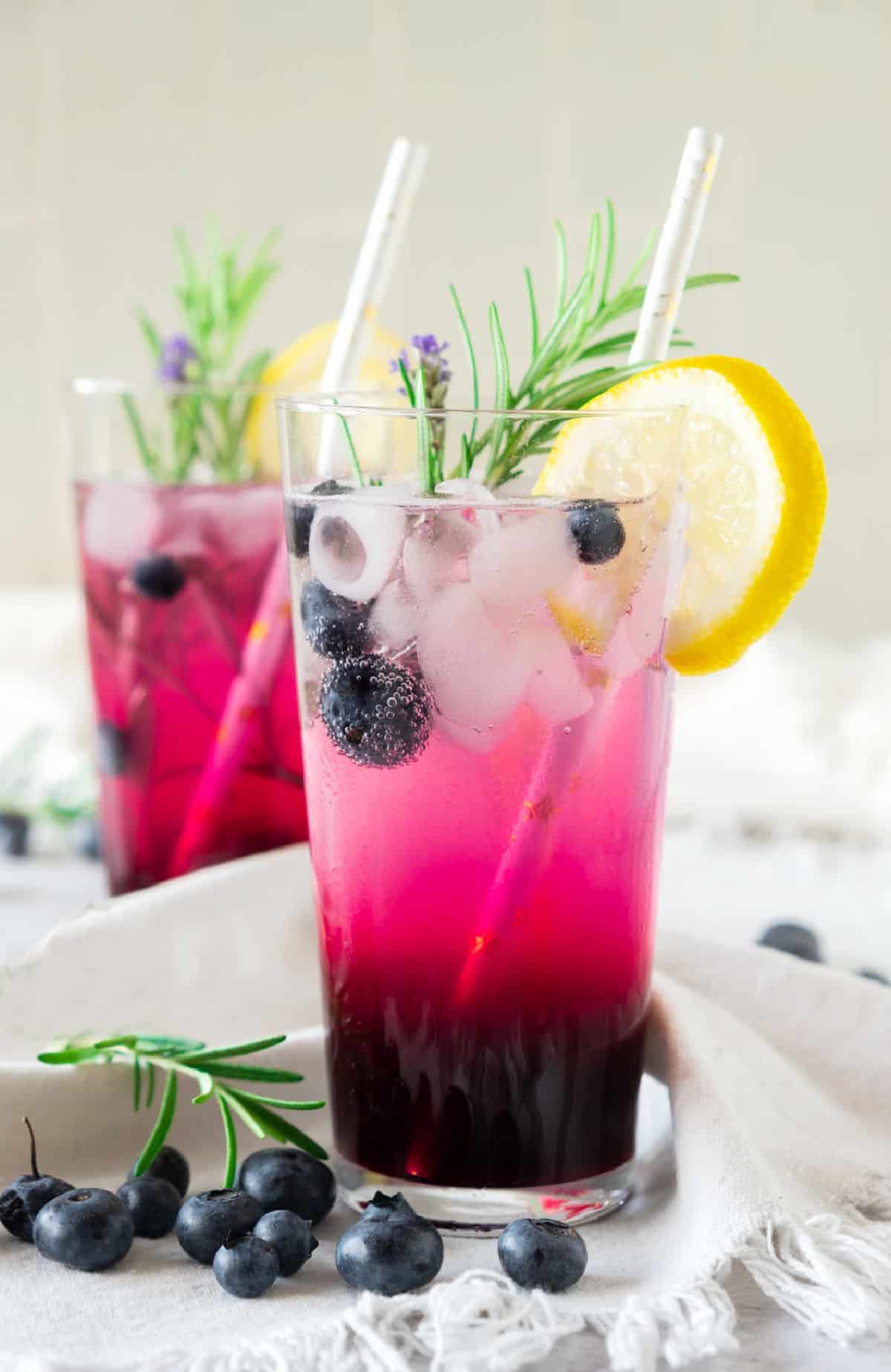 Glasses with blueberry lemonade with lemon slices and rosemary sprigs. White surface, beige background.