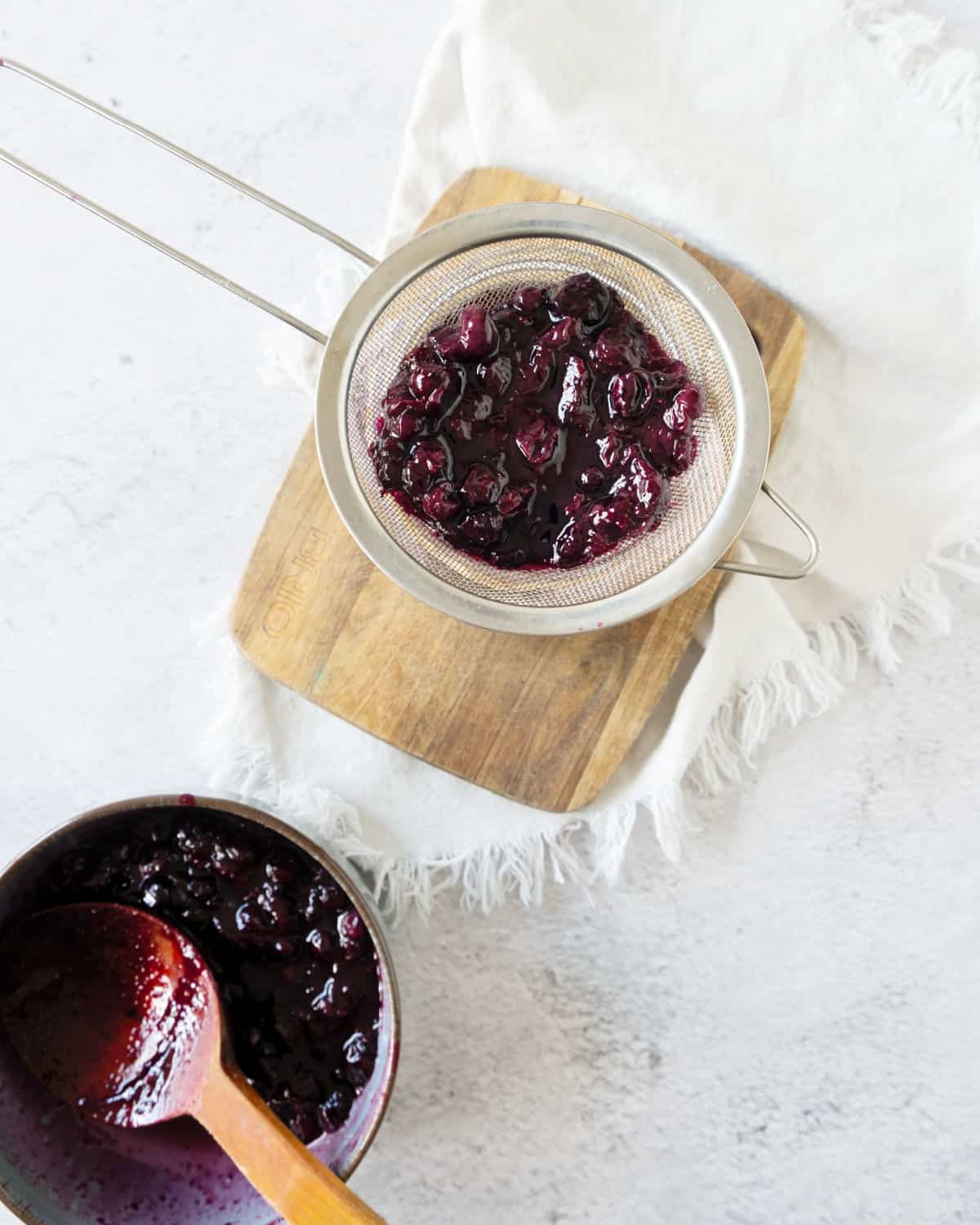 White surface with blueberry syrup in saucepan, some on a colander on a wooden board.