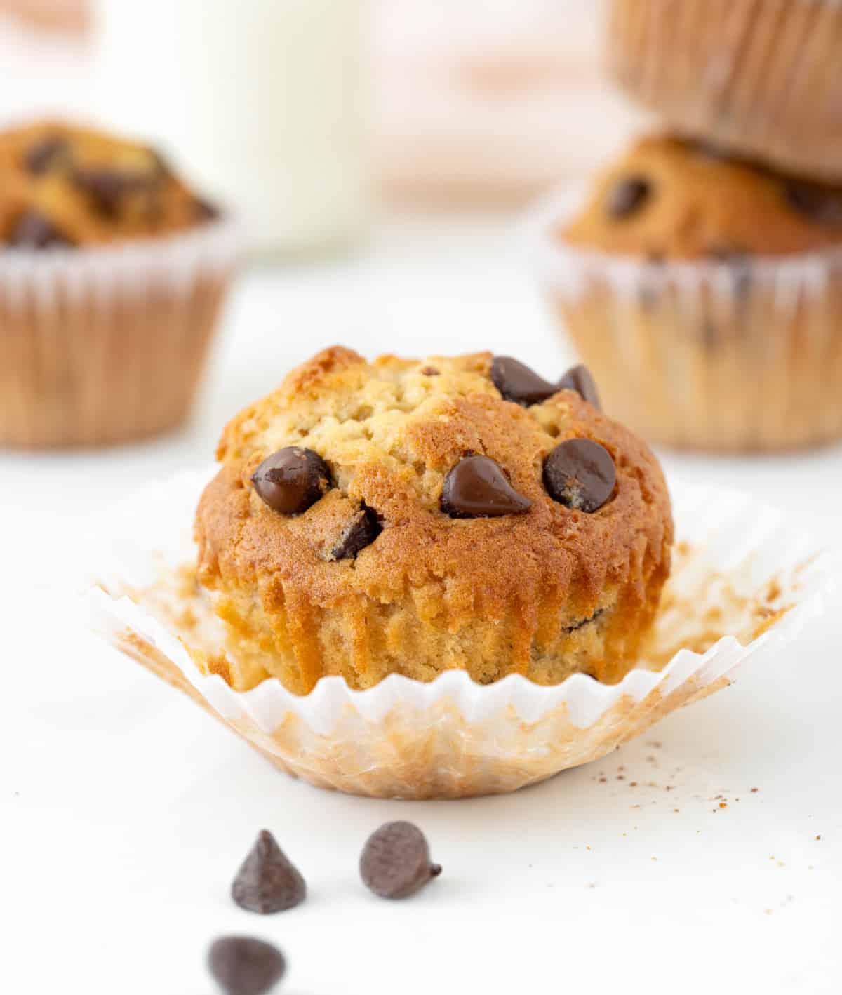 Opened paper liner with chocolate chip muffin on a white surface. More muffins in the background.