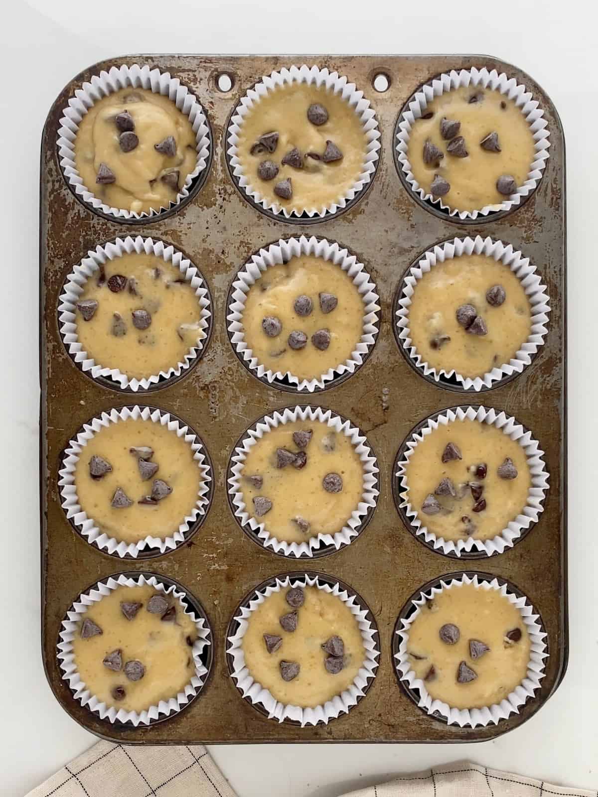 Chocolate chip muffin batter in a metal pan with paper liners. White surface.