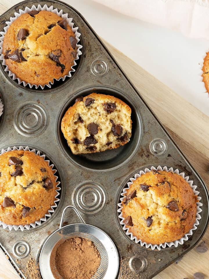 Vintage metal pan with chocolate chip muffins, whole and halved. Light wooden board, white surface.
