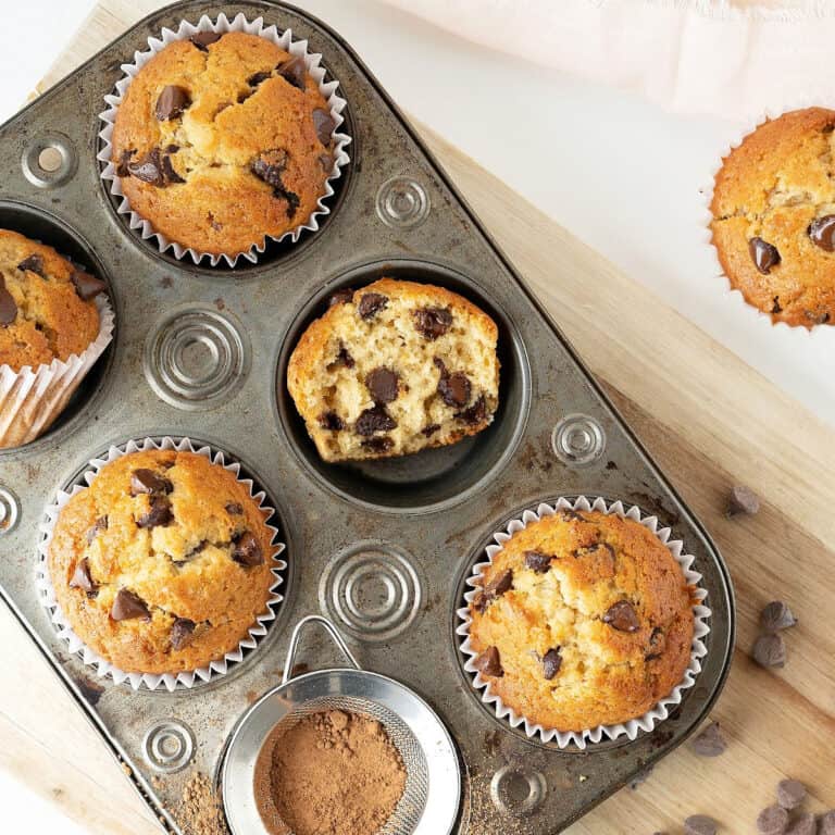 Vintage metal pan with chocolate chip muffins, whole and halved. Light wooden board, white surface.