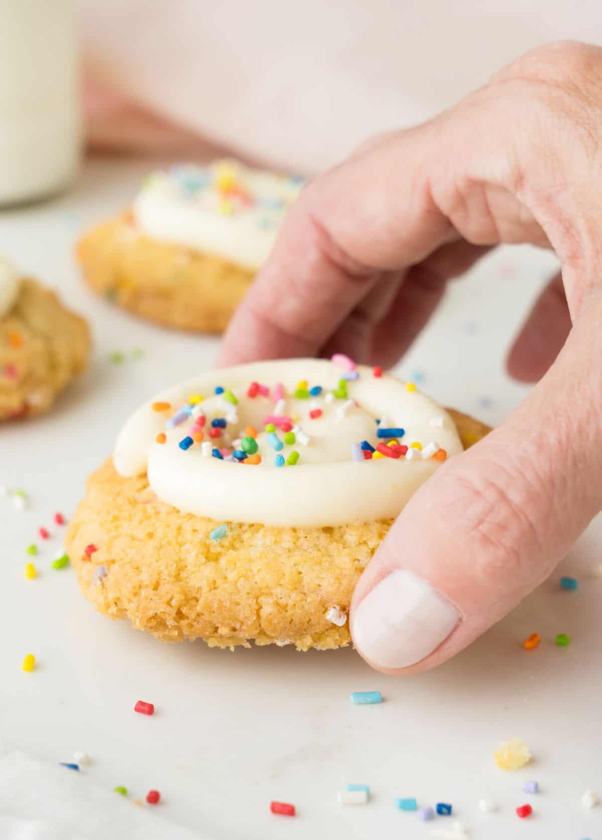 Lifting a frosted funfetti cake cookie. White surface, pink background, more cookies. 