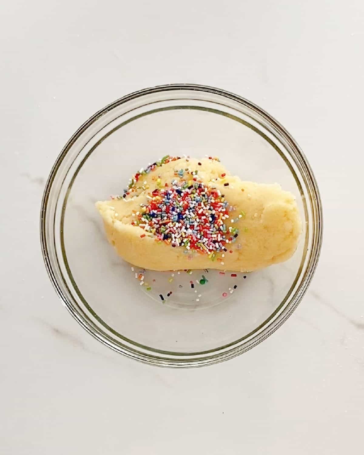 Sprinkles added to cookie dough in a glass bowl on a white surface.