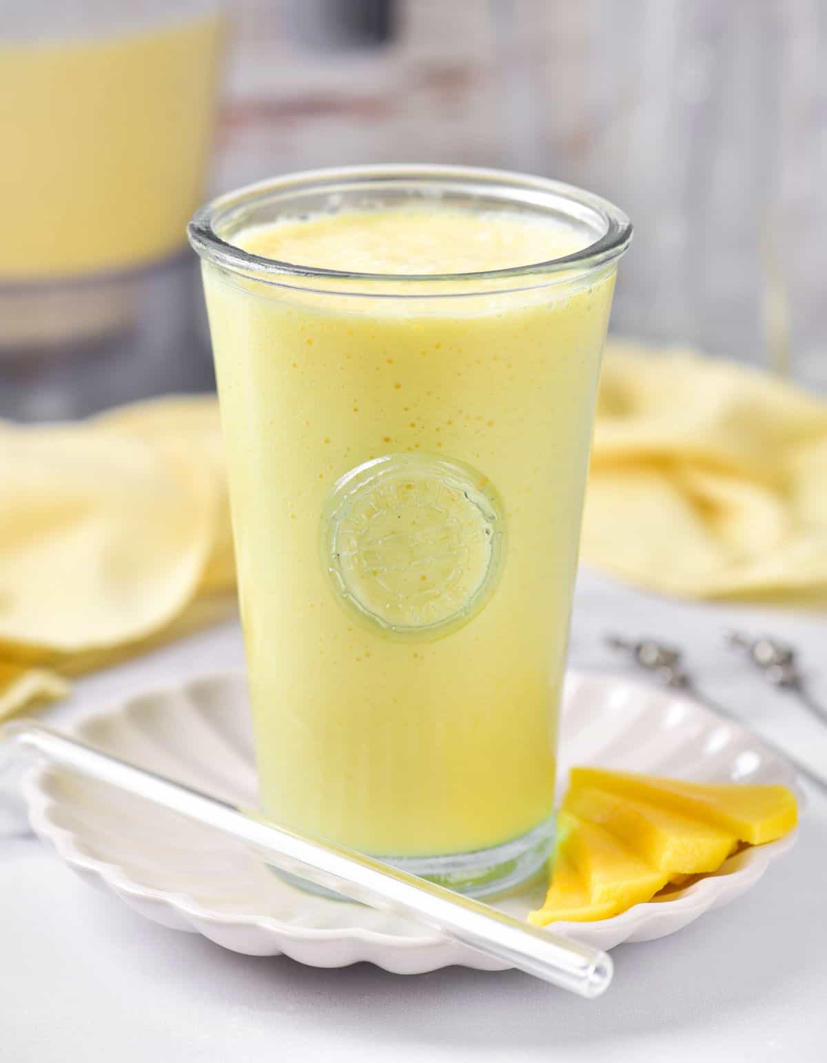 Mango milkshake in a tall glass on a white plate. Grey and yellow background, white surface. 
