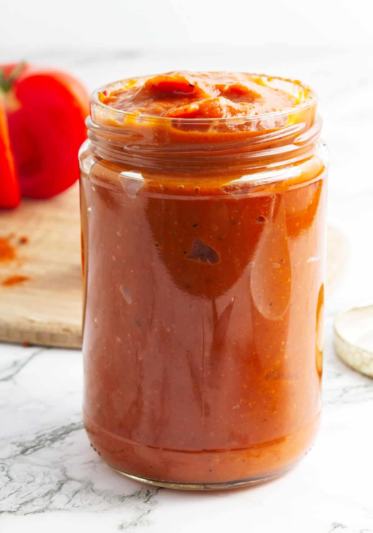 Jar of homemade ketchup on a white marbled surface. Tomato and wooden board in the background.