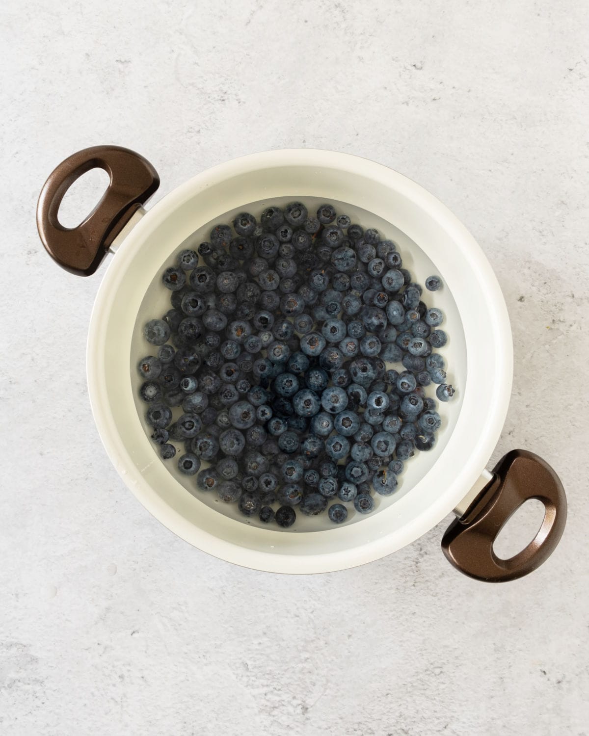 Saucepan with blueberries and simple sugar syrup. Light grey surface. 