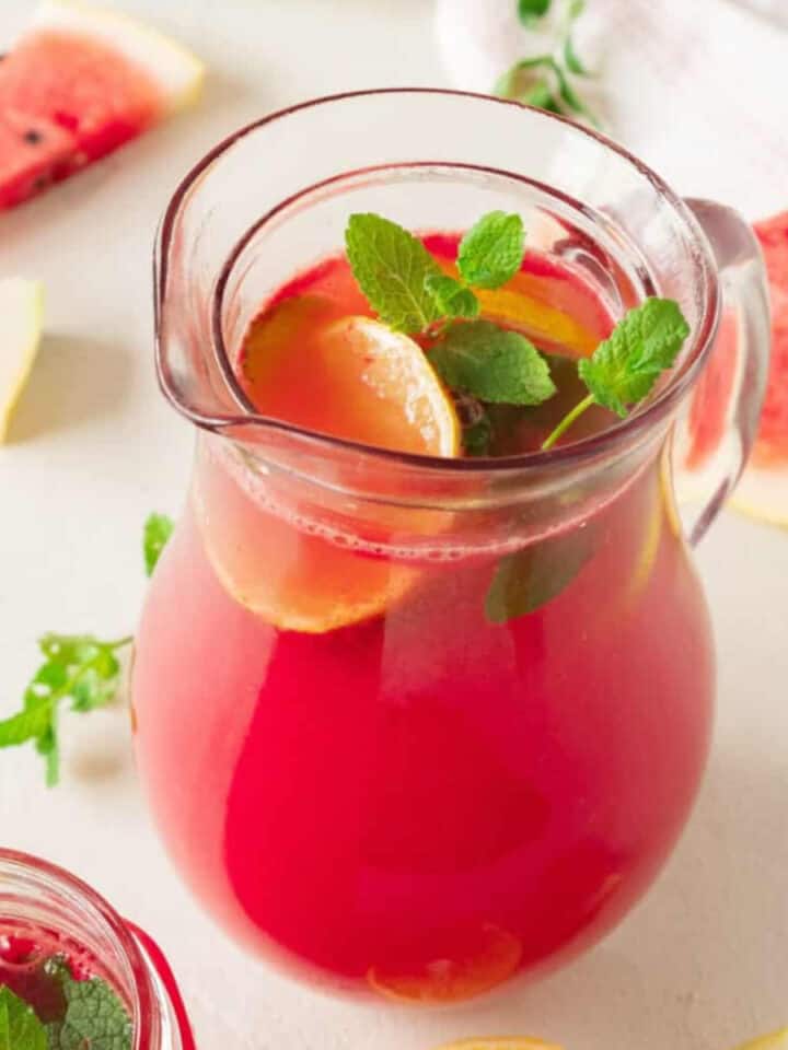 Off-white surface with pitcher of watermelon lemonade with mint sprigs. Watermelon slices around.