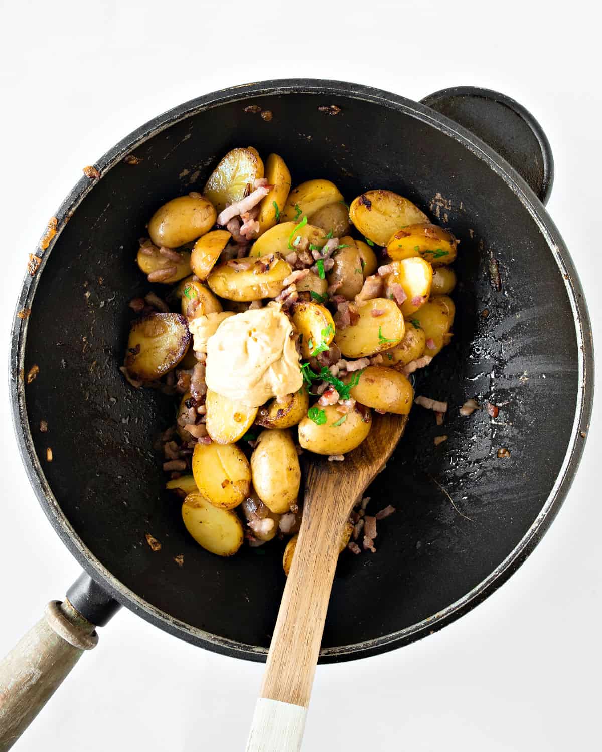 Halved baby potatoes, bacon and mustard in a black skillet. White background.